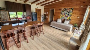 Une chambre confortable, aménagée dans le style d’un chalet, avec un plancher en bois, une kitchenette, un coin repas doté de tabourets de bar, un lit orné d’une fresque colorée et une fenêtre donnant sur un jardin.