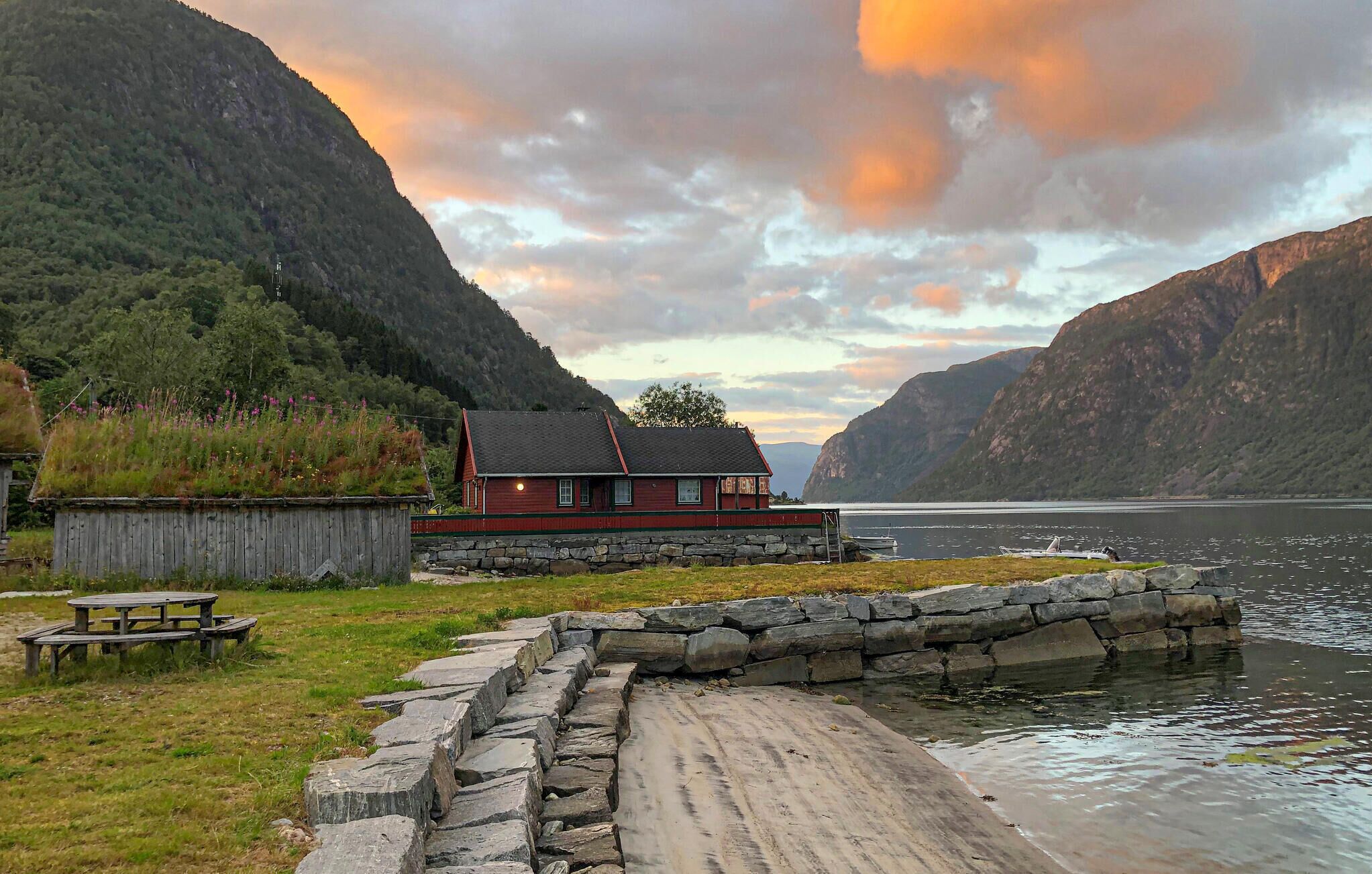 Uma cabana rústica com base de pedra, deck de madeira e vista para um lago rodeado por montanhas.