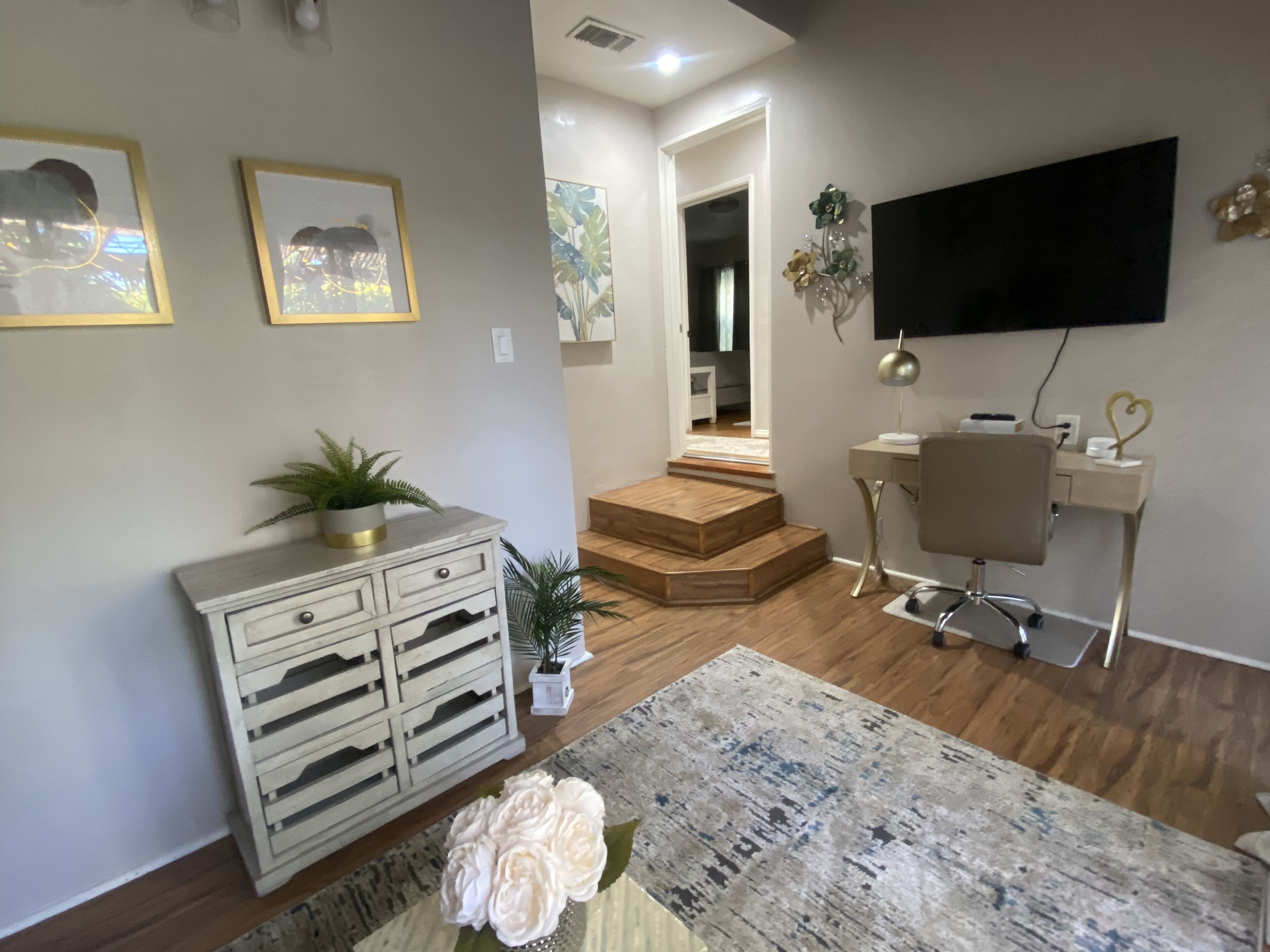 A living room with a sofa, a coffee table, a TV mounted on the wall, a desk with a chair, and a staircase leading to another room.
