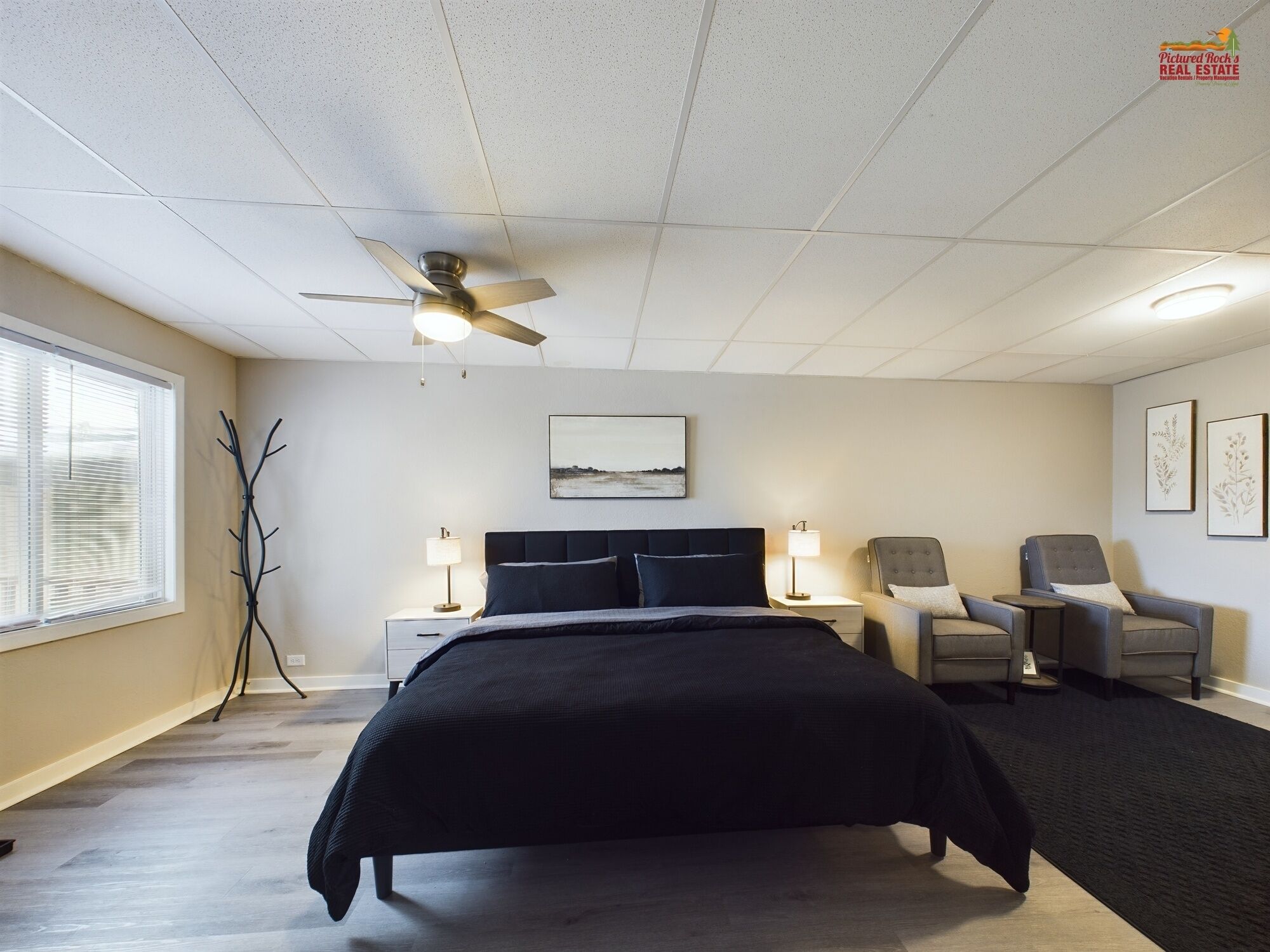 A bedroom with a bed, two bedside tables, a chair, a standing lamp, a ceiling fan, and a window with blinds.