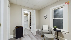 Un couloir avec une porte donnant sur un placard, un fauteuil gris, un petit pouf et une fenêtre avec une plante posée sur une table d’appoint.