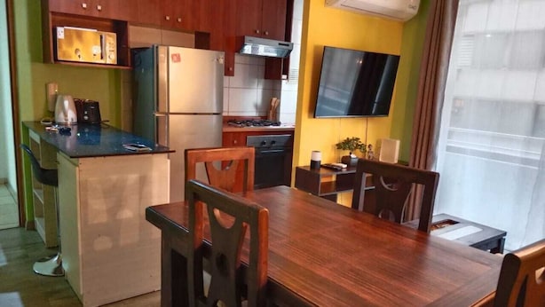 A compact kitchen with a refrigerator, microwave, and stove. There is a dining table with chairs and a TV mounted on the wall. - Apart Latitud (Santiago)