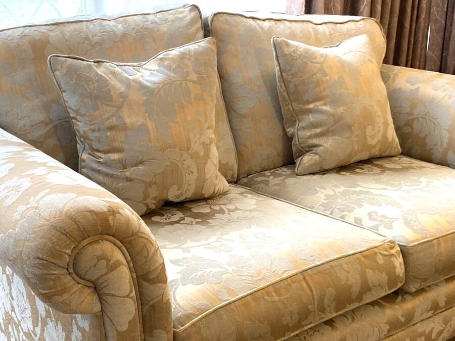 A beige upholstered sofa with a floral pattern, featuring a tufted armrest and three cushions.