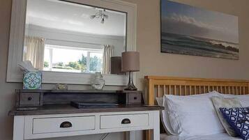 A bedroom with a bed, a bedside table with a lamp, a dresser with drawers, a mirror, and a painting of a seascape.