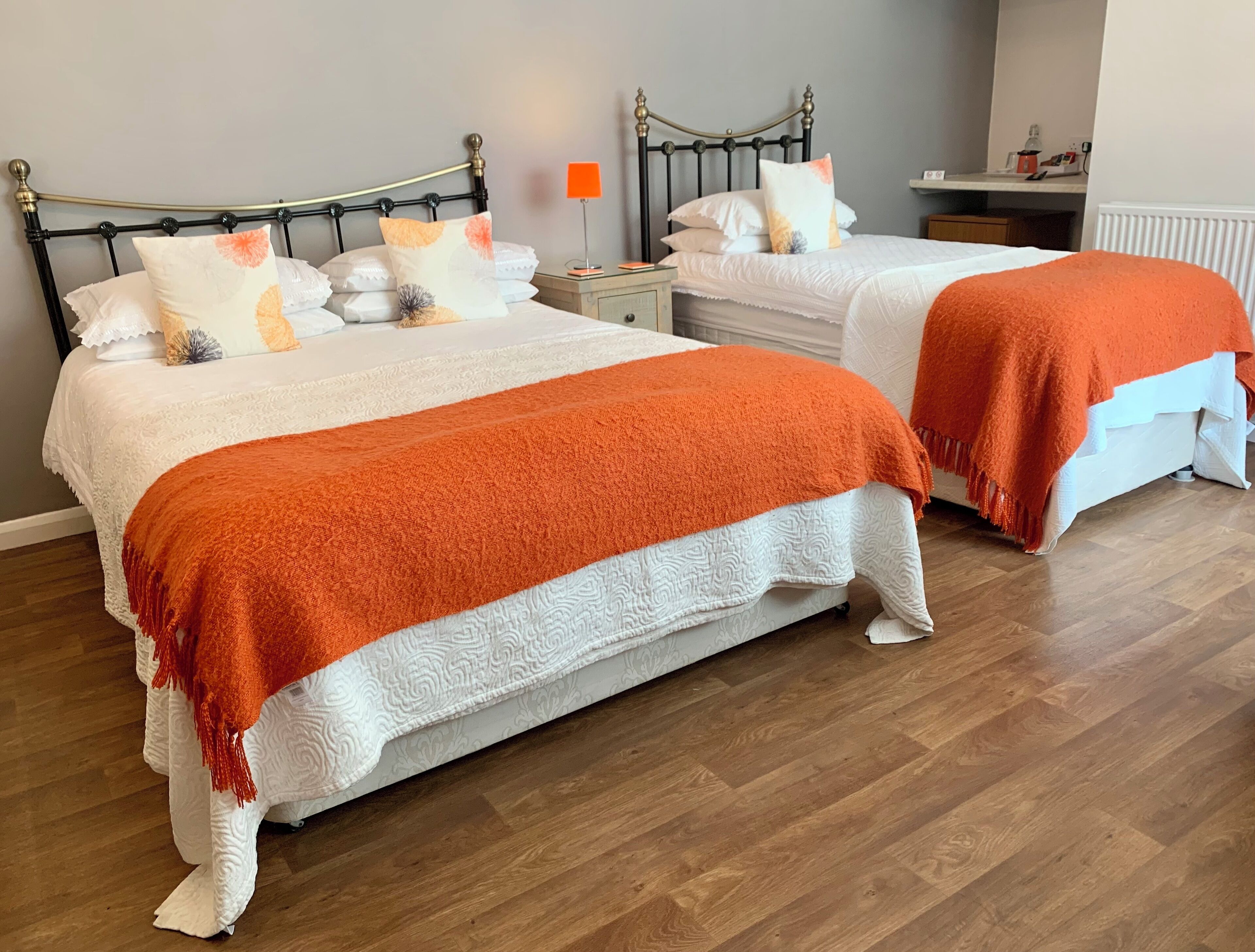 A double bed with an orange blanket and white bedding, a nightstand with a lamp, and a bedside table with a red lamp.