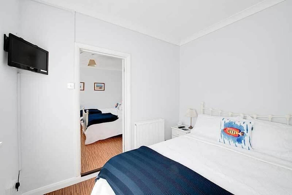Quarto de hotel com uma cama, televisão na parede, mesa de cabeceira com abajur e uma porta que dá para outro quarto.