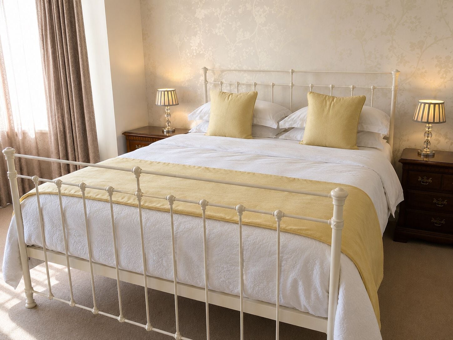 Una cama bien tendida con estructura metálica blanca, dos mesitas de noche con lámparas y un estampado floral en la pared.
