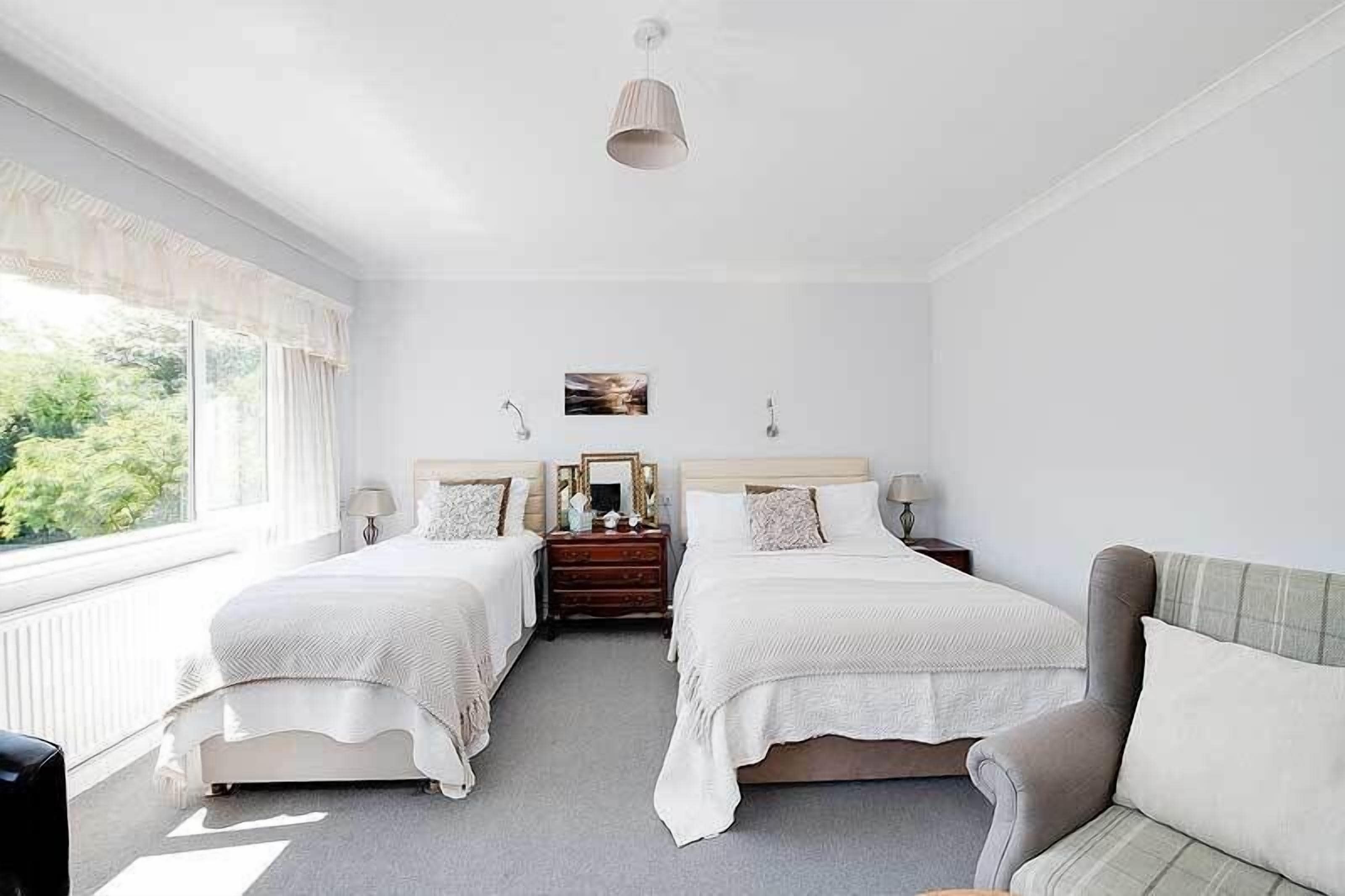 A bedroom with two beds, a dresser, a mirror, and a window with a view of greenery.