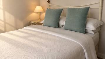 Un letto con una coperta bianca e strutturata, due cuscini, un comodino con una lampada e una testiera bianca.