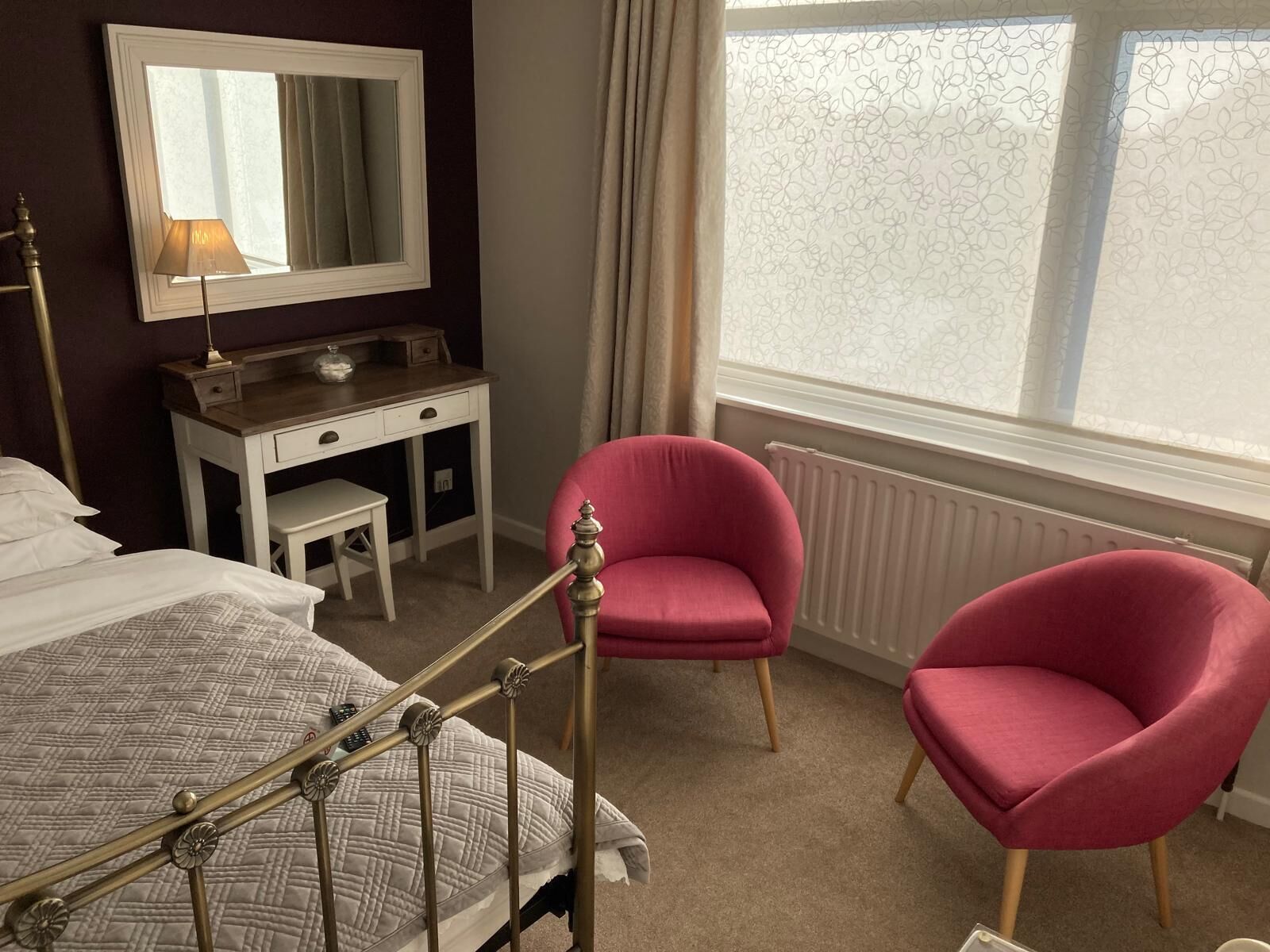 A hotel room with a bed, a desk, two pink chairs, a mirror, and a window with curtains.