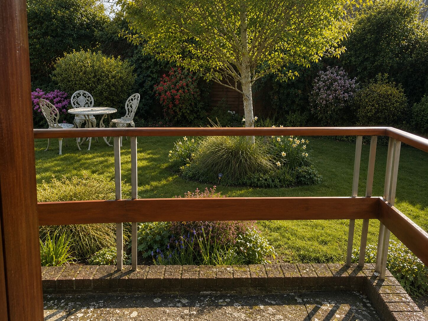 Un balcón de madera con barandilla de metal da a un jardín con césped, una mesa pequeña y dos sillas.
