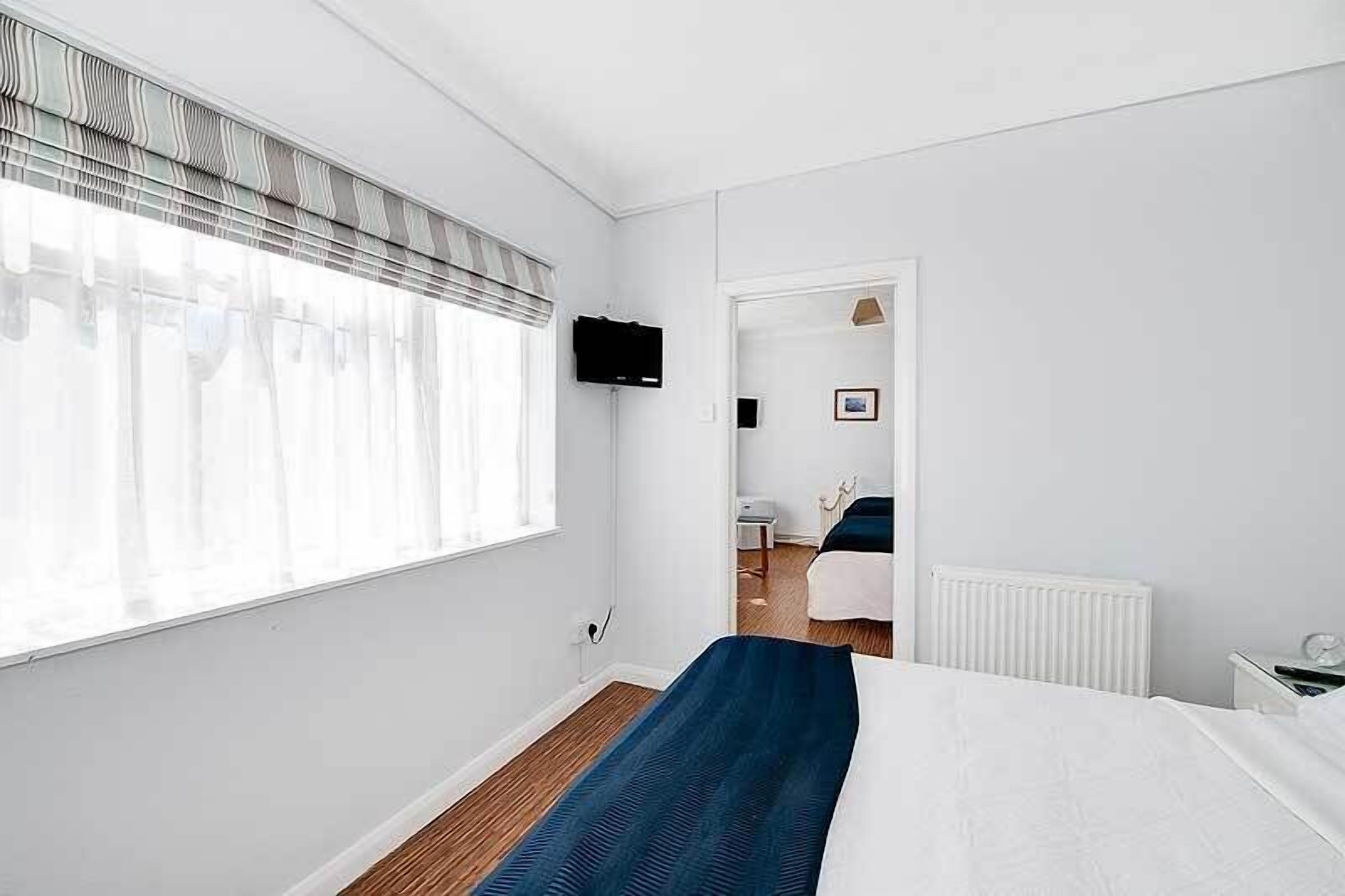 Une chambre d’hôtel avec une grande fenêtre, un lit recouvert d’une courtepointe bleue, un radiateur blanc et un téléviseur fixé au mur.