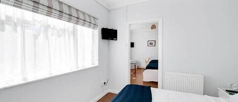 Une chambre d’hôtel avec une grande fenêtre, un lit recouvert d’une courtepointe bleue, un radiateur blanc et un téléviseur fixé au mur.