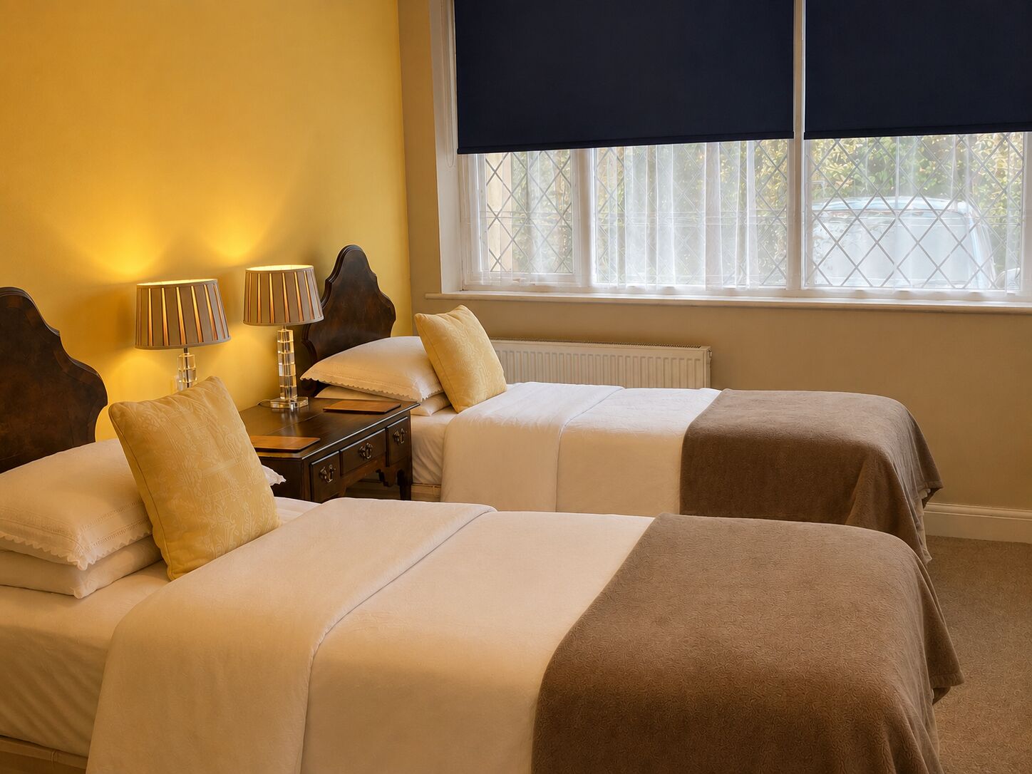 A hotel room with two beds, a nightstand, lamps, and a window with blinds.