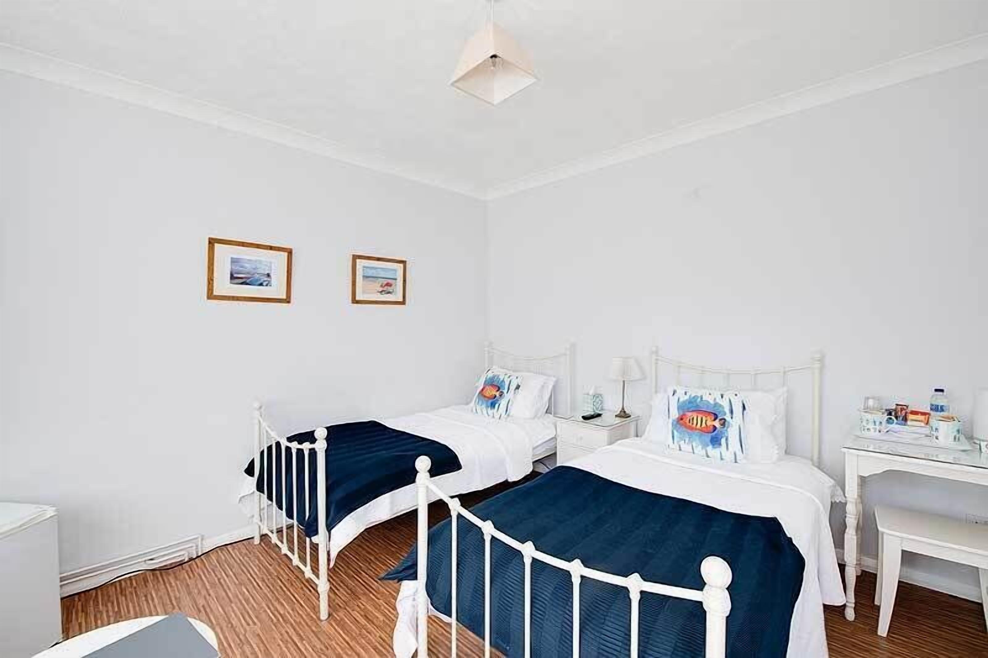 A room with two single beds, white walls, and a wooden floor. There are two framed pictures on the wall, a white desk with a lamp, and a small table with a bottle and a cup.