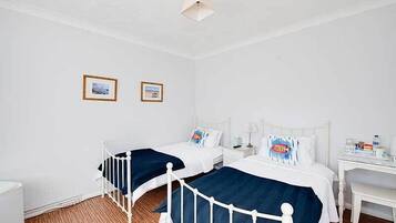 A room with two single beds, white walls, and a wooden floor. There are two framed pictures on the wall, a white desk with a lamp, and a small table with a bottle and a cup.