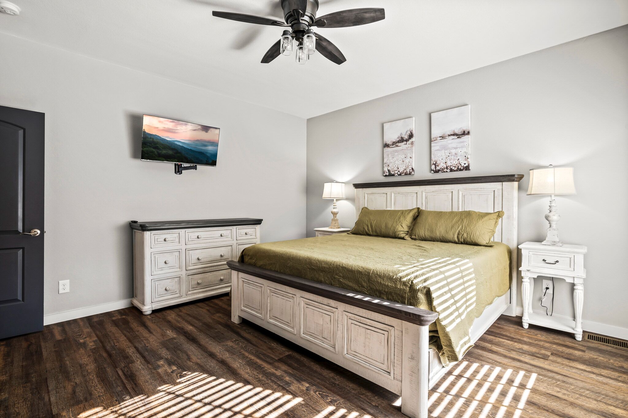Une chambre à coucher comprenant un lit, des tables de chevet, un ventilateur de plafond, un téléviseur et des œuvres d’art accrochées au mur.