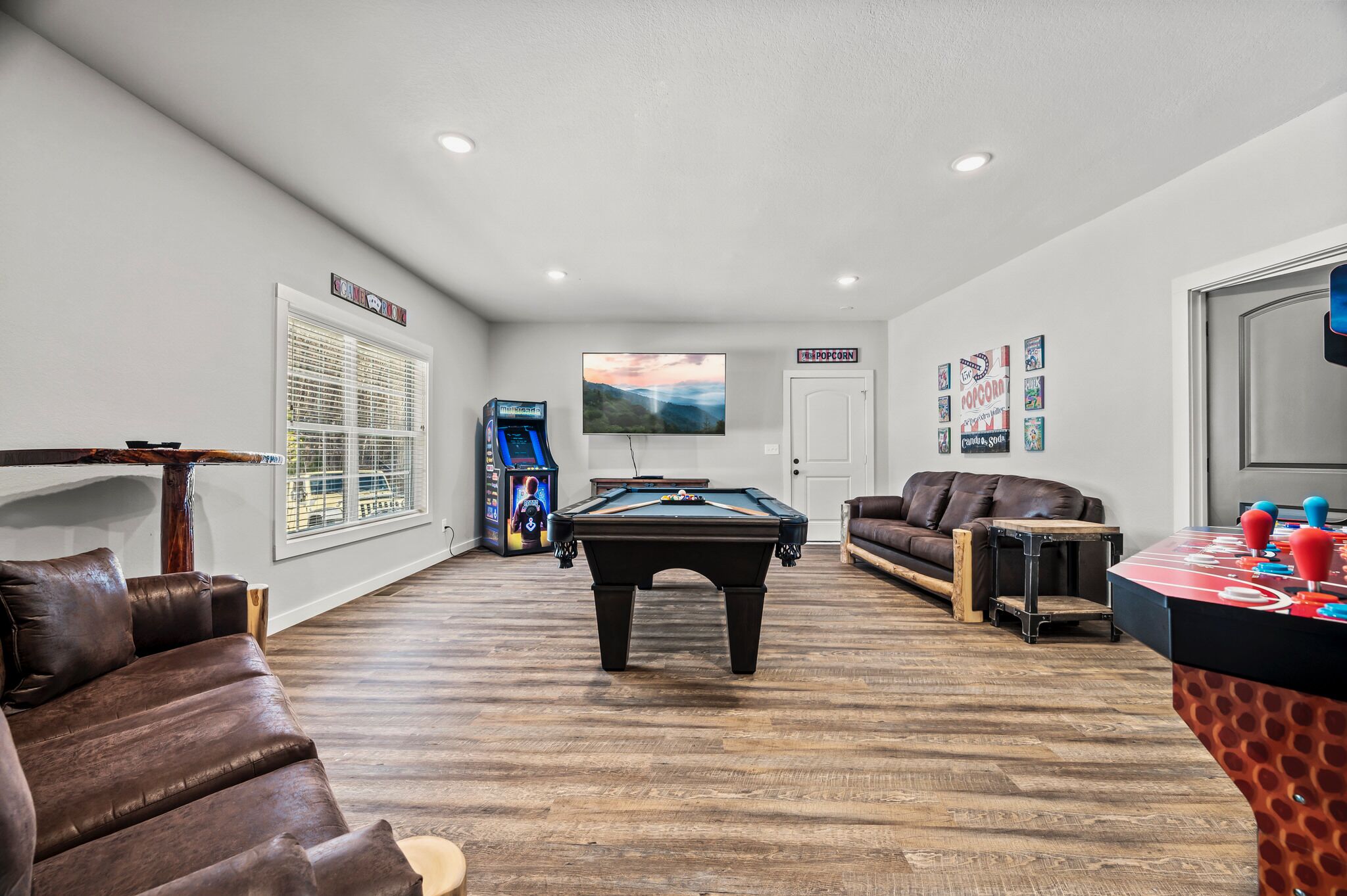 A room with a pool table, a sofa, a foosball table, and a television.