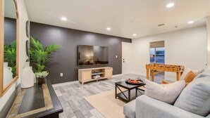 A modern living room with a flat-screen TV, a wooden entertainment unit, a grey sofa, and a foosball table. - Best of Midtown-3 Bedrooms, Minutes to Attractions (Houston)