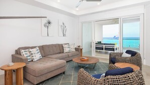A modern living room with a sofa, coffee table, and wicker chairs, a ceiling fan, and a view of the ocean through large sliding doors. - Rum Point Resort #305 by Grand Cayman Villas (Seven Mile Beach)