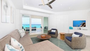 A modern living room with a sofa, two armchairs, a coffee table, and a television. There is a ceiling fan and a view of the ocean through large glass doors. - Rum Point Resort #305 by Grand Cayman Villas (Seven Mile Beach)