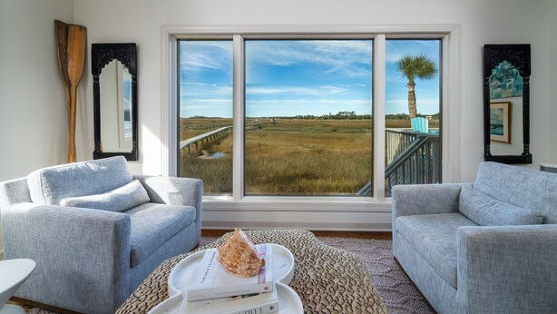 A room with two grey sofas, a wooden paddle, a large window with a view of a field and palm tree, and a framed artwork on the wall.