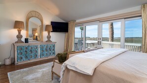 A bedroom with a large bed, a dresser, a television, and a balcony with a view of the landscape.