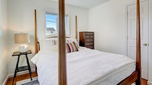 A four-poster bed with white bedding, a wooden nightstand with a lamp, a dresser with drawers, and a window with blinds. - Mystic Island Bay Breeze, Beautiful waterfront sunsets waiting (Little Egg Harbor Township)