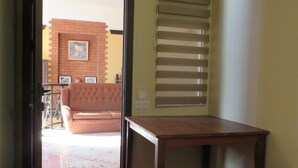 A hotel room with a bed, a wooden table, a window with blinds, a door leading to a balcony, and a tiled floor.