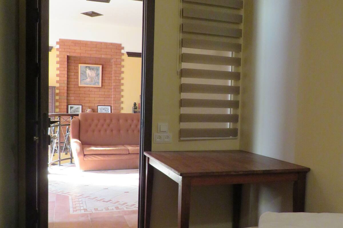 Habitación de hotel con cama, una mesa de madera, una ventana con persianas, una puerta que da a un balcón y piso de baldosas.