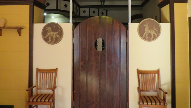 A room with a wooden door, two wooden chairs, a patterned rug, and a ceiling light fixture.