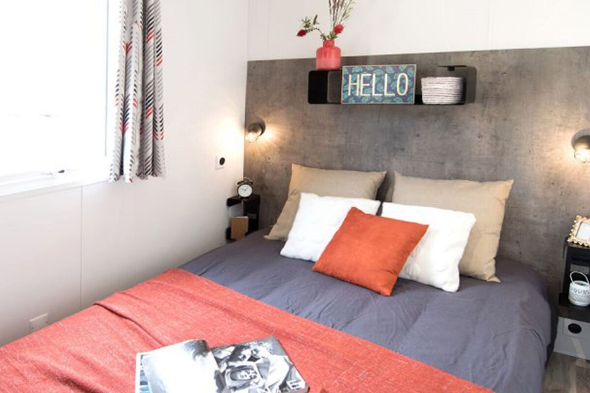 A modern bedroom with a bed, a nightstand, a wall clock, a 'HELLO' sign, and a window with curtains.
