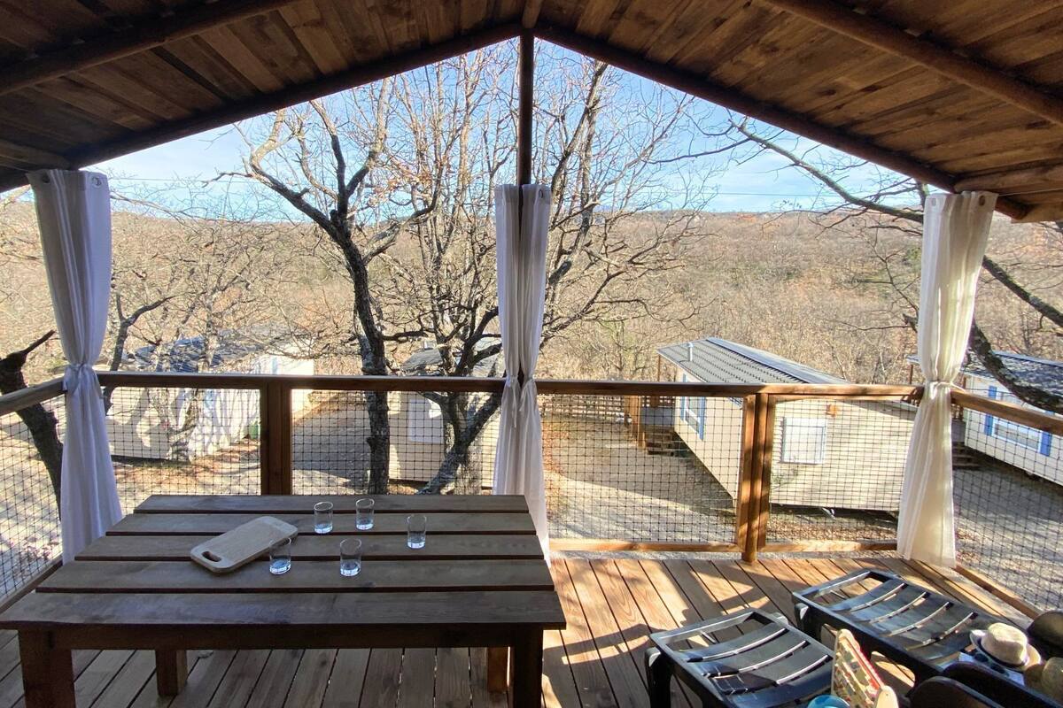 Una terraza de madera con una mesa y sillas, con vistas a un paisaje pintoresco con árboles y un edificio distante.
