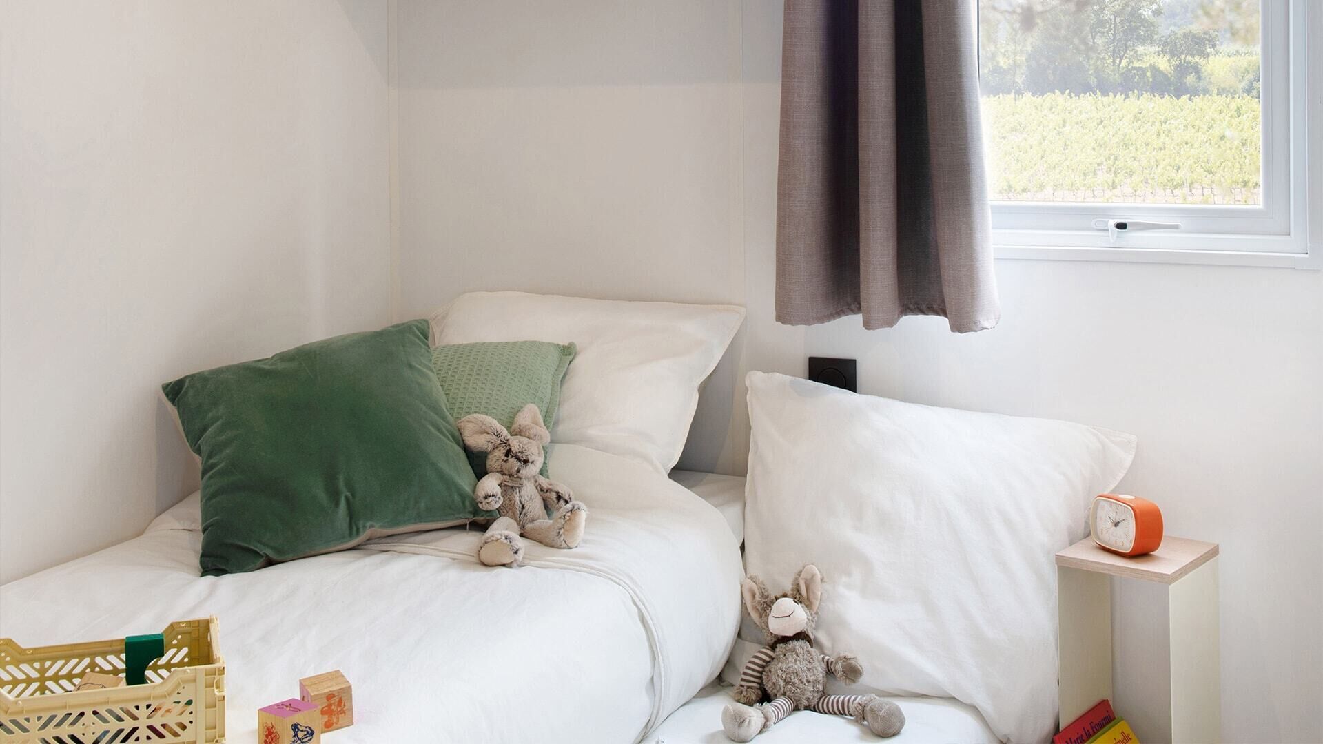 A bedroom with a bed, pillows, stuffed toys, a bedside table with a clock, and a window with curtains.