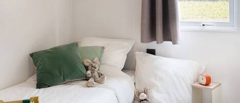 A bedroom with a bed, pillows, stuffed toys, a bedside table with a clock, and a window with curtains.
