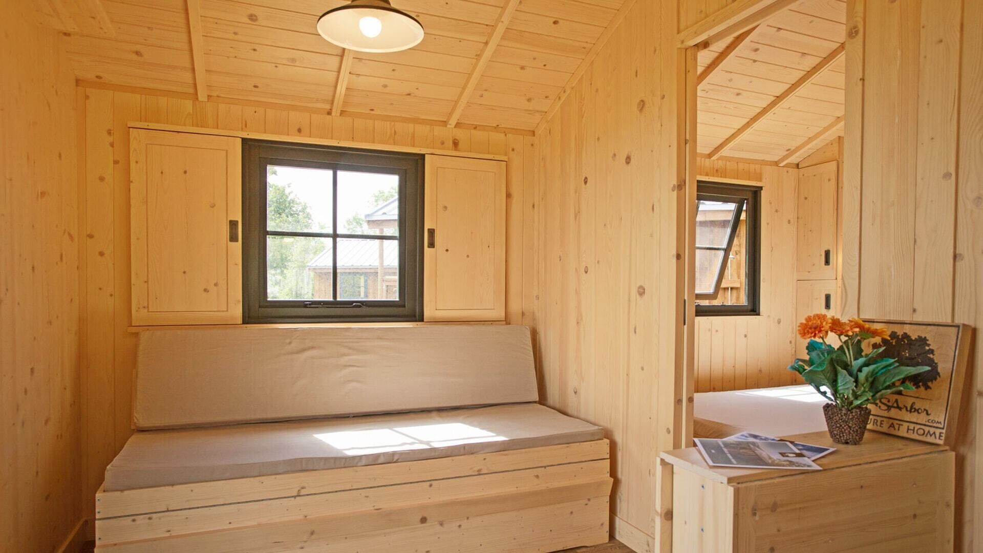 A wooden cabin interior with a bench, a window, and a small table with a plant.