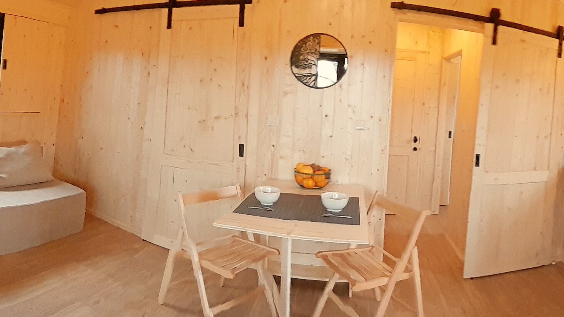 A cozy room with a wooden interior, a round mirror on the wall, a table with two chairs, and a bowl of fruit on the table.
