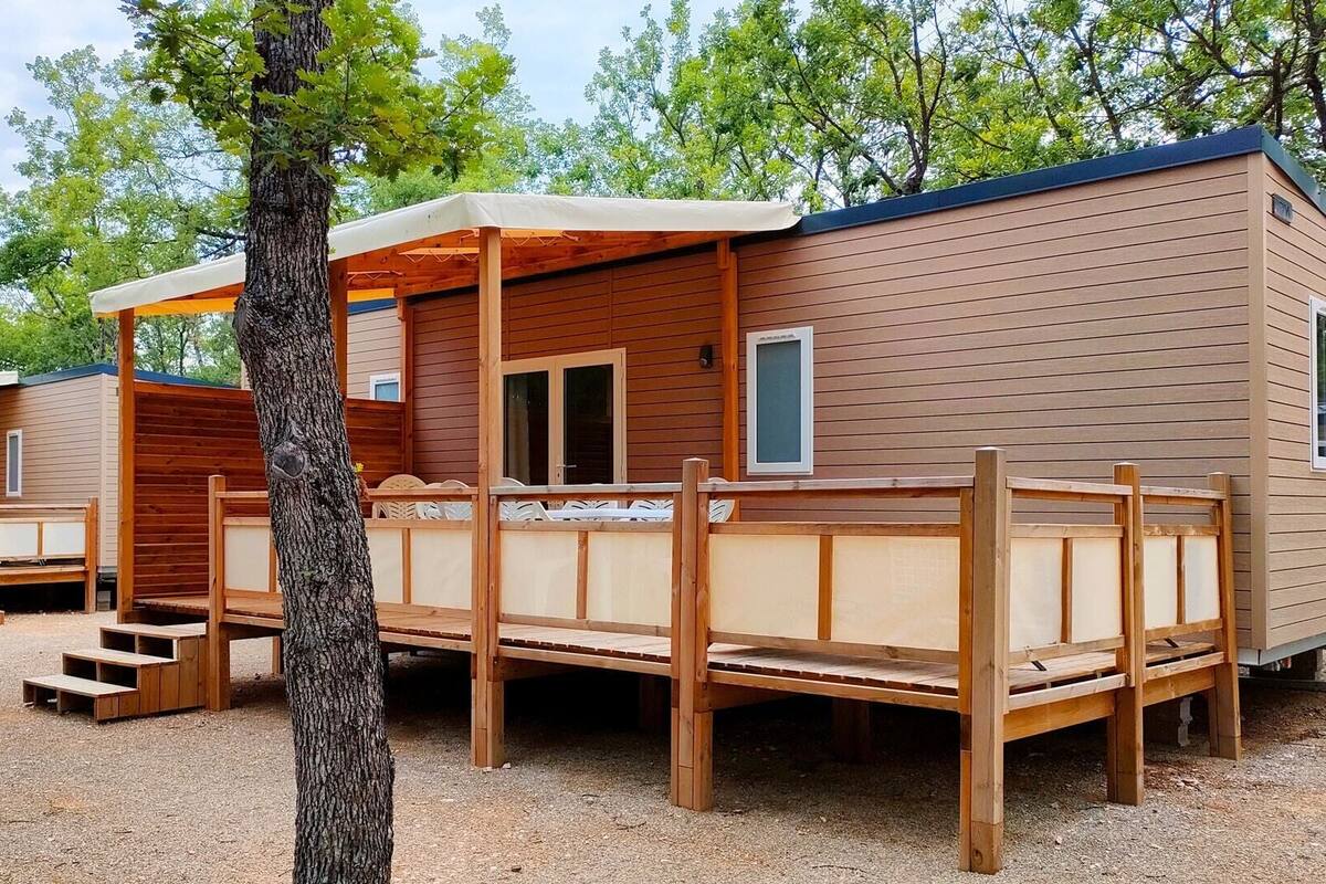 Una hilera de cabañas de madera con terraza y un espacio cubierto, rodeadas de árboles.