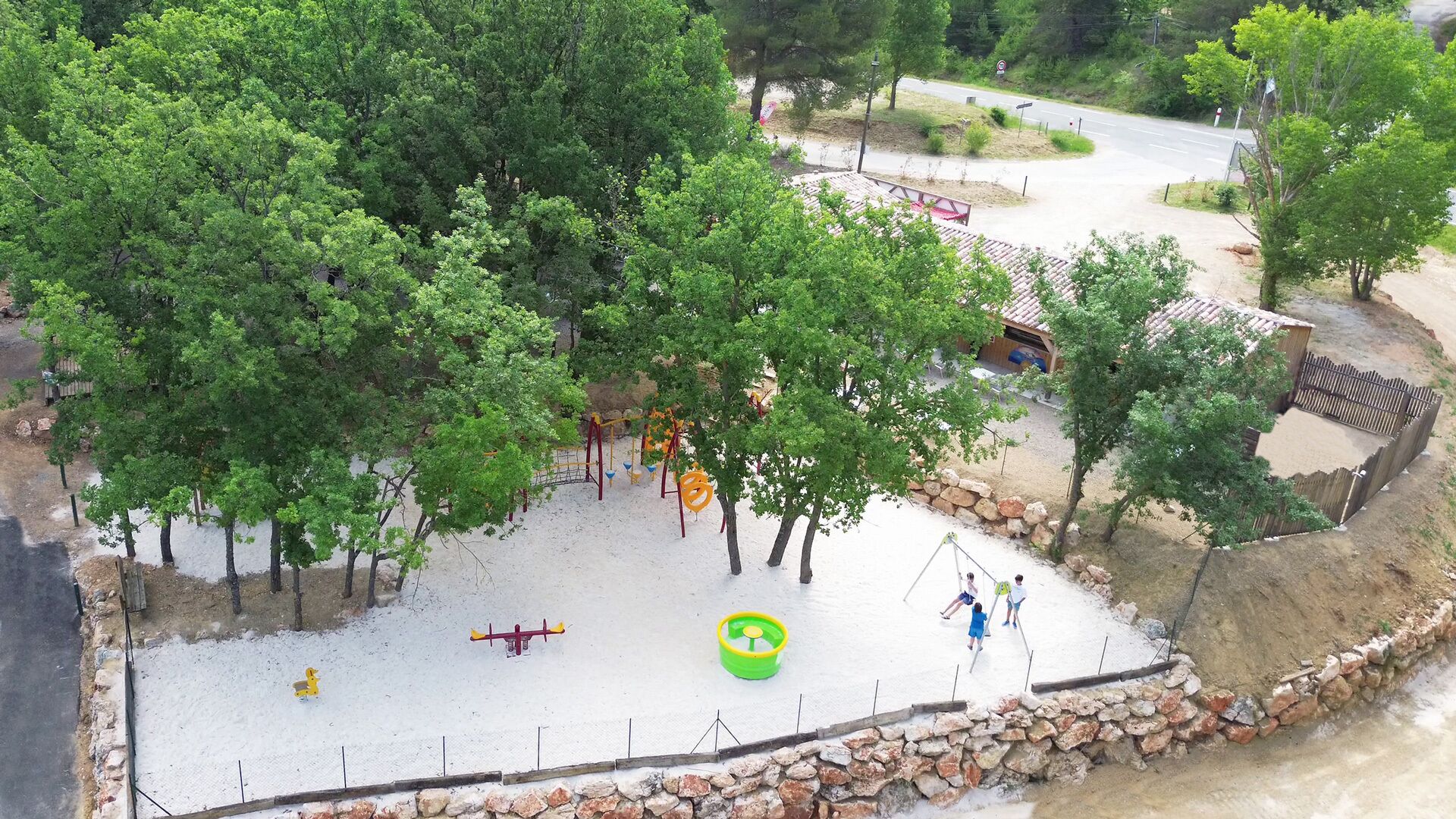 Zona arenosa para juegos con columpios y un tobogán, rodeada de árboles y un muro de piedra.