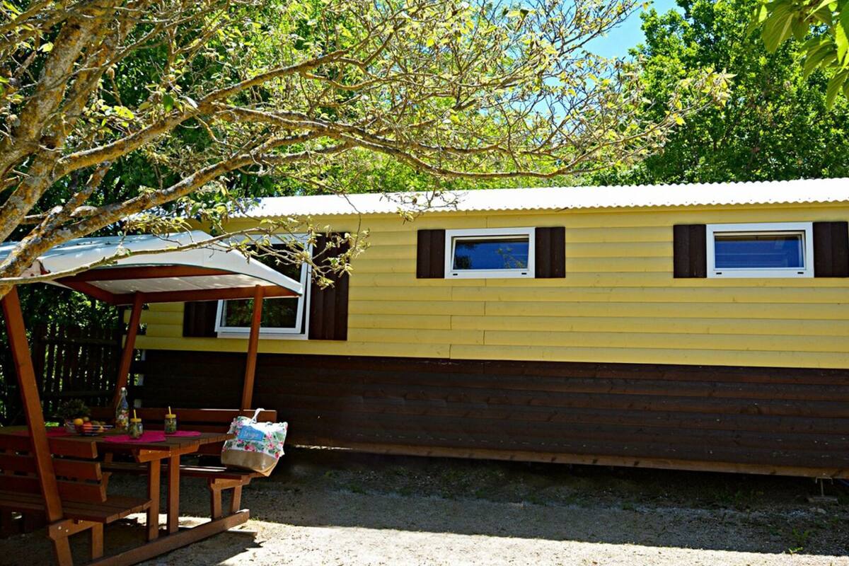 Una casa móvil amarilla y marrón con dos ventanas, un espacio de estar techado al aire libre y una mesa con sillas.