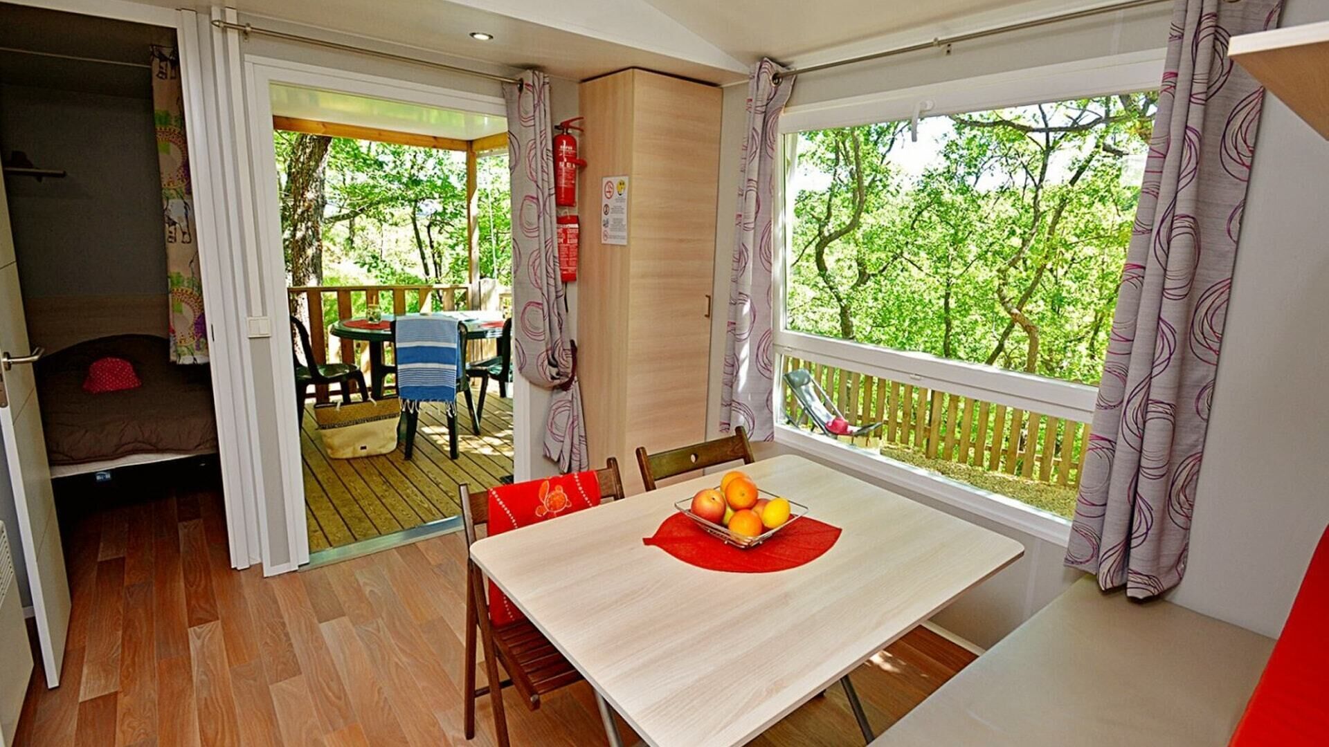 A room with a wooden floor, a dining table with a red placemat, and a window with a view of trees.