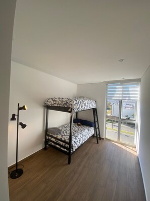 A bunk bed with a patterned bedspread, a standing floor lamp, and a large window with blinds.