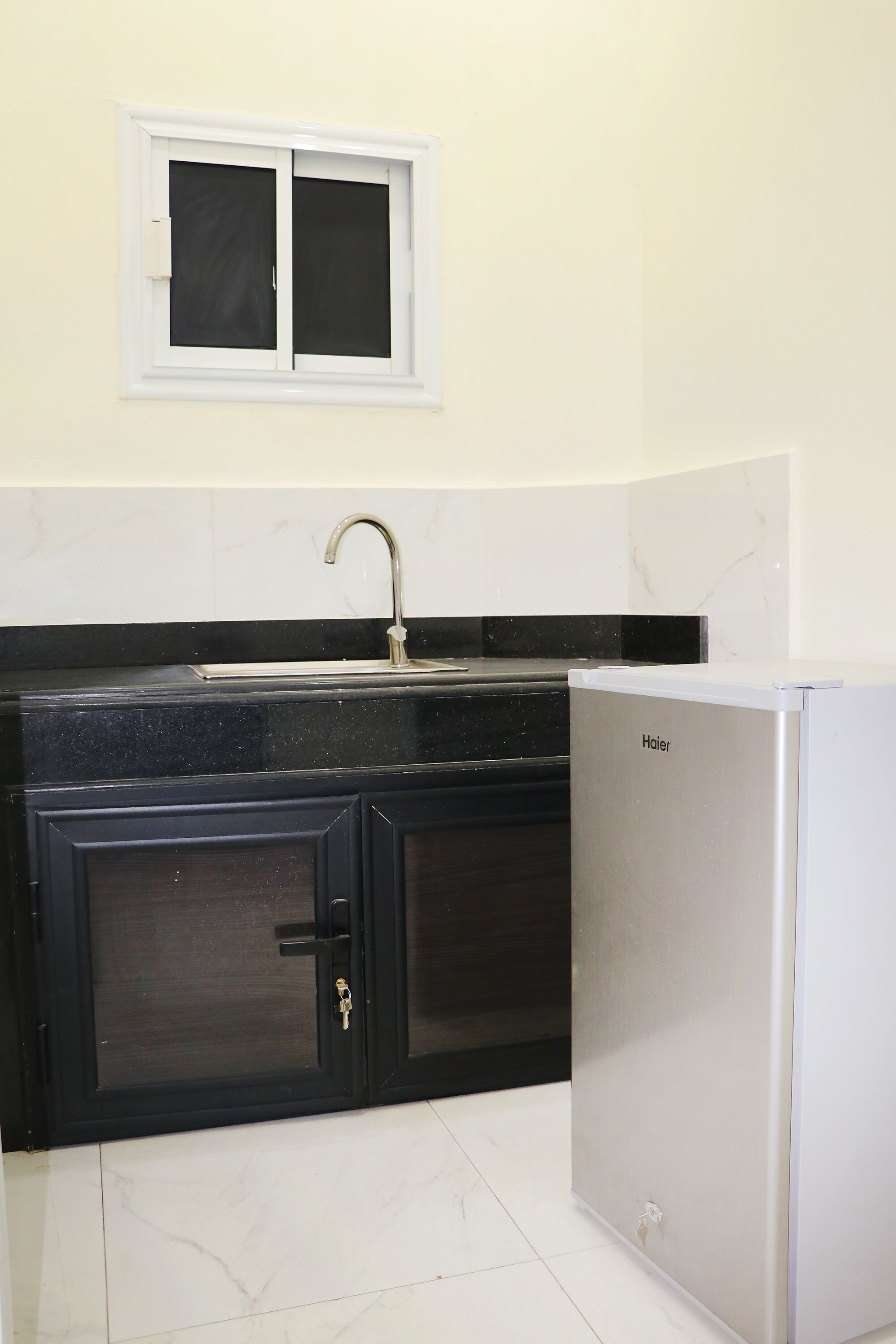Una cocina compacta con encimera negra, un lavamanos blanco, un refrigerador Haier y una ventana con marco blanco.
