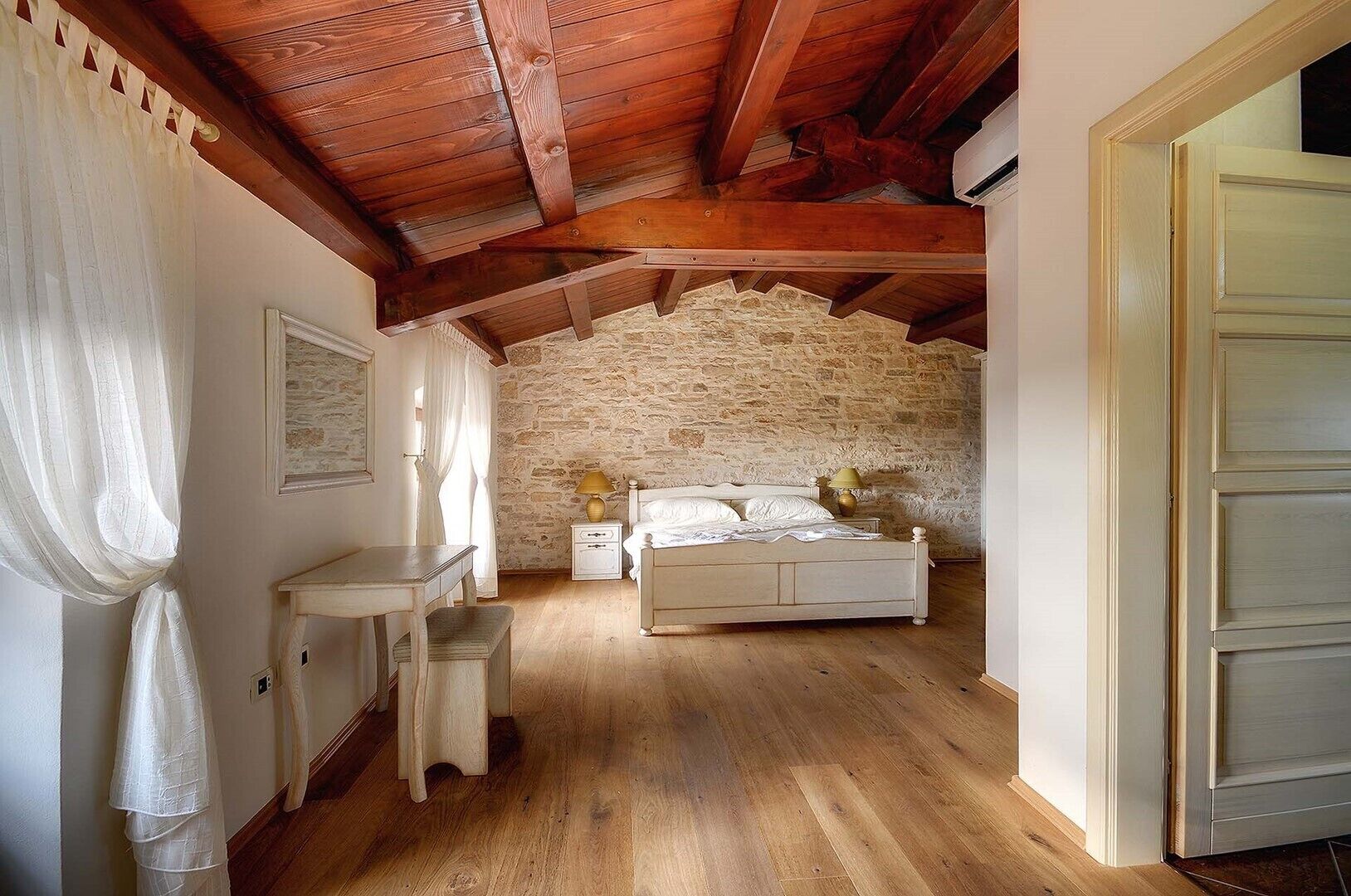 Une chambre avec un mur en pierre, des poutres en bois au plafond, un lit, des tables de chevet et un bureau.