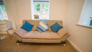 Un sofá color beige con cojines azules en una habitación con una ventana, una pequeña mesa y una silla.