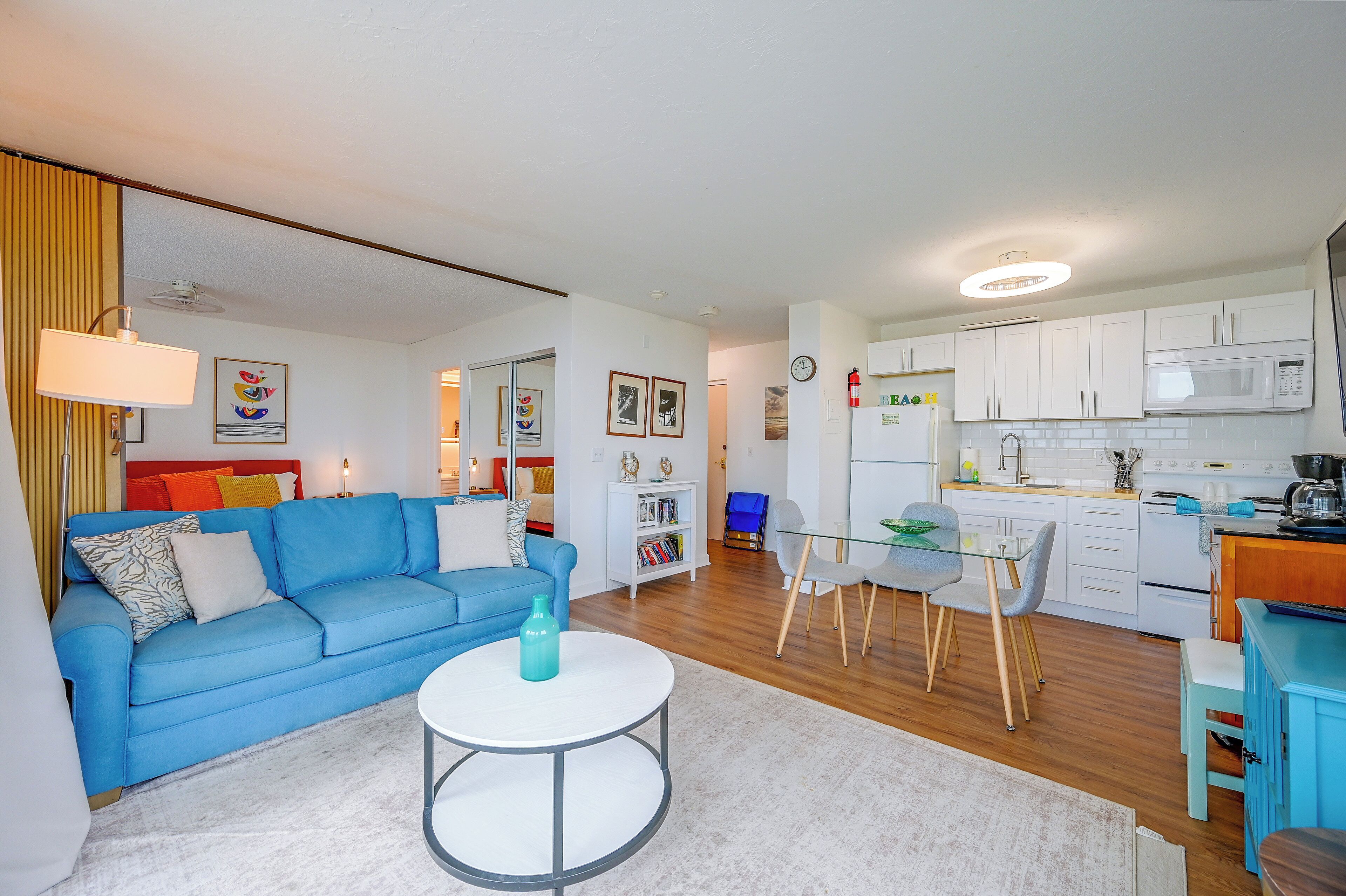 Un apartamento moderno con un sofá azul, una mesa redonda, una cocina con armarios blancos y una zona de comedor.