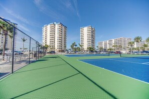 Una cancha de tenis con una cerca de malla metálica, rodeada de edificios altos y palmeras.