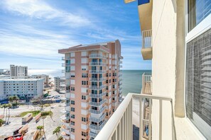A high-rise hotel with multiple balconies, a view of the beach, and a parking area with palm trees.
