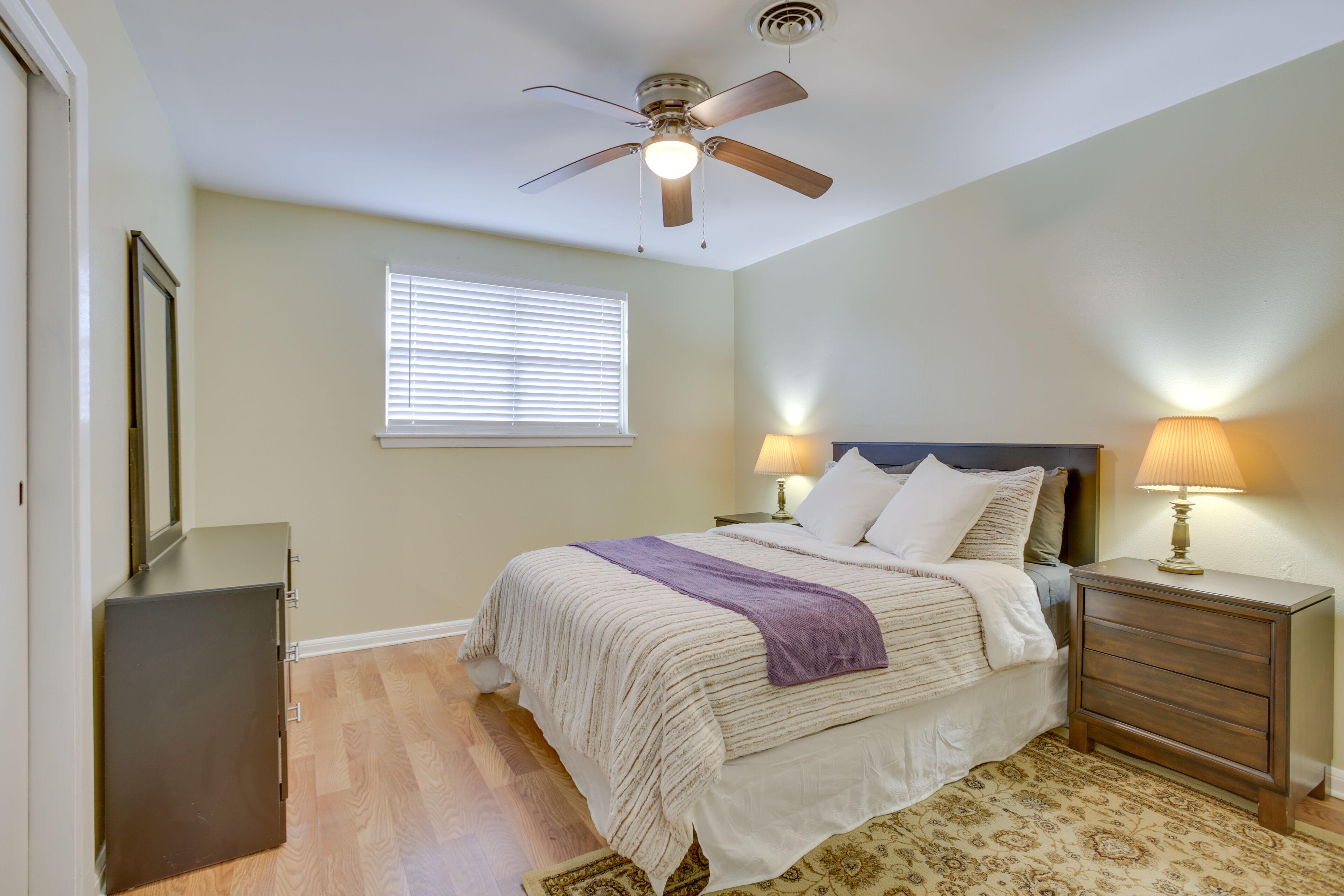 A bedroom with a bed, bedside lamps, a ceiling fan, a window with blinds, a dresser, and a patterned rug.