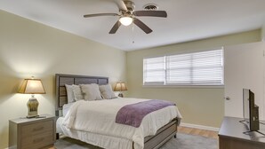 Un dormitorio con cama, lámparas de noche, ventilador de techo, ventana con persianas, escritorio y televisor.